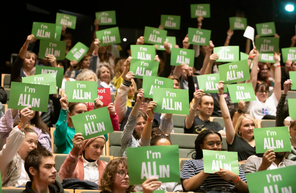 An audience of people holding green postcards in the air that says "I'm All In".