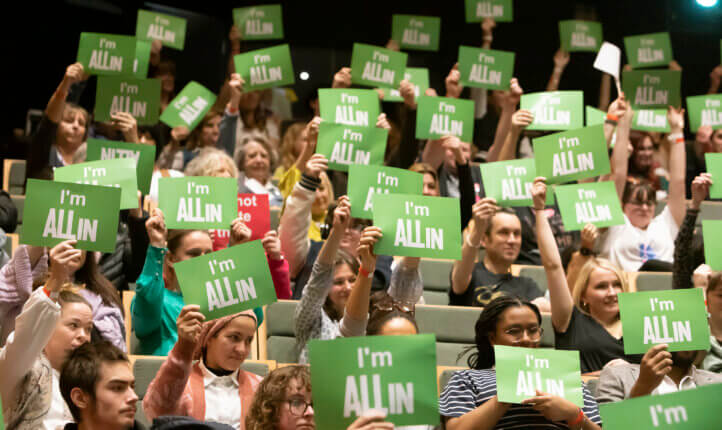 An audience of people holding green postcards in the air that says "I'm All In".