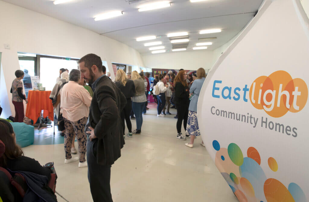 People stood together at the All In Festival 2023. An Eastlight Community Homes sign is situated to the right of the image.