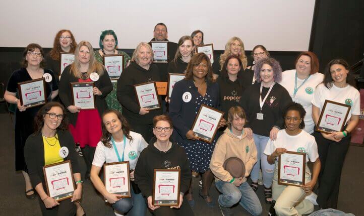 • A group of All In programme participants holding their certificates stating 'Community Entrepreneur on the first All In Community Incubator 2022-2023', smiling at the camera. Behind them is a screen with the hashtag #WeAreAllIn and the Easltight Community Homes Logo