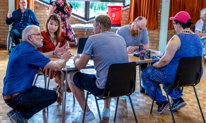 A group of people are sat around a table. There is a man crouched down infront of the table talking to a man and woman who are sat at the table.